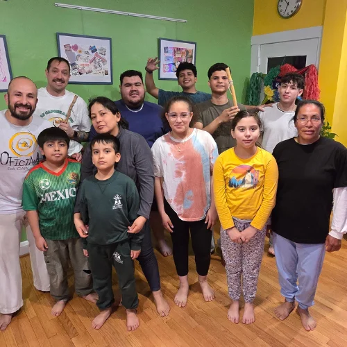 Group of 14 smiling students and instructors in a martial arts studio, barefoot on a wooden floor with green walls and framed artwork in the background.