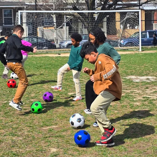 Group of kids and an adult coach practicing soccer drills on a grassy field, with colorful balls lined up and a goal in the background.