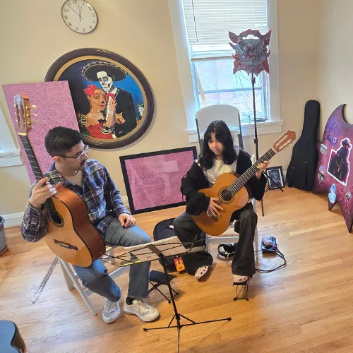 Two guitar players sit on chairs in a bright room, each with an acoustic guitar; a music stand with sheet music sits between them.