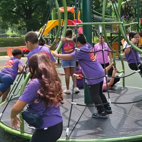 Children in purple shirts climb on a circular jungle gym at a sunny park playground.