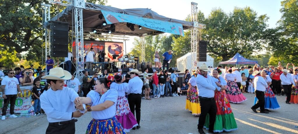 Couples in colorful traditional Mexican dresses and white shirts dance in front of a stage at an outdoor festival.
