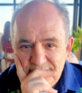 Close-up of an older man with gray hair resting his chin on his hand, wearing a dark blue shirt, with a soft background blur