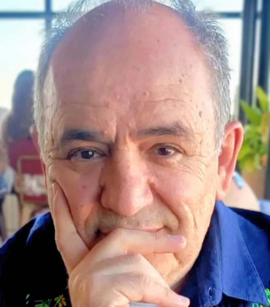 Close-up of an older man with gray hair resting his chin on his hand, wearing a dark blue shirt, with a soft background blur