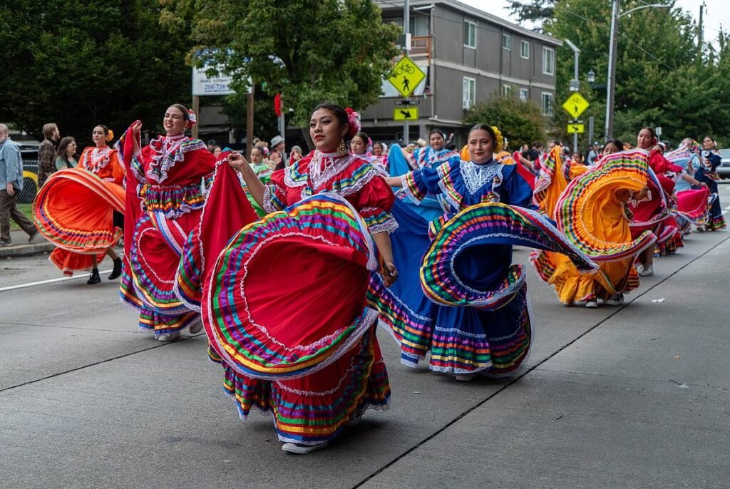 Group of women in bright multicolored Mexican folkloric dresses dancing in a street parade.