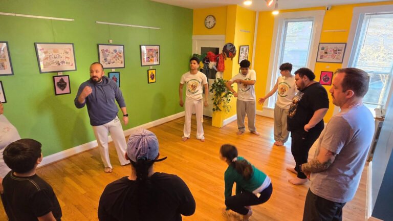 Instructor leads a small group in a martial arts or movement class in a bright green and yellow studio, students standing around watching a demonstration with one arm raised.