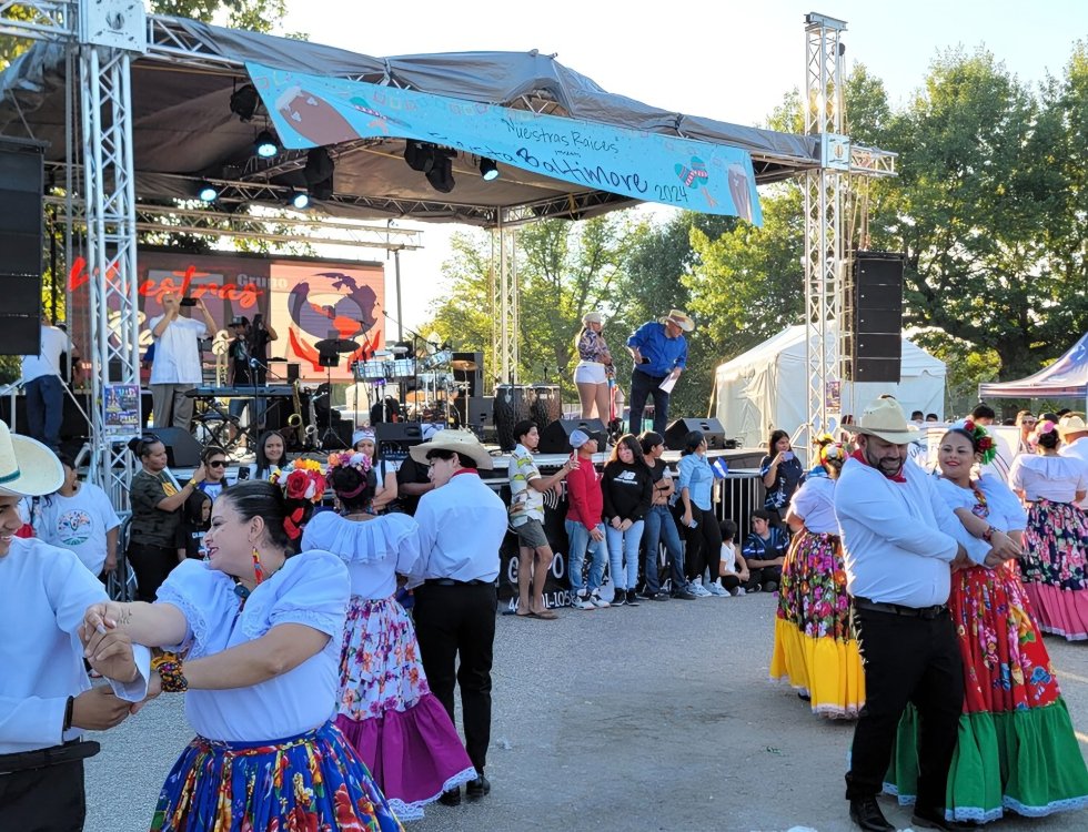 Dancers in bright floral skirts perform folkloric dances in front of a stage with a live band at an outdoor festival.