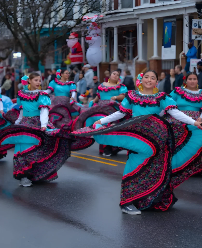 Group of dancers in turquoise folklorico dresses with pink/black embroidery performing a street parade.