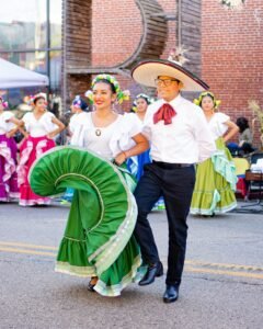 Couple in folklorico attire dancing in the street; woman wears a white blouse and green skirt, man in white shirt with a large sombrero.