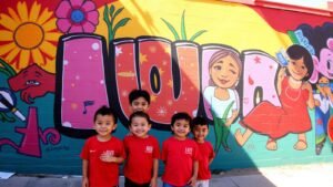 Five smiling children in red shirts pose in front of a colorful mural with large letters, flowers, and cartoon characters.