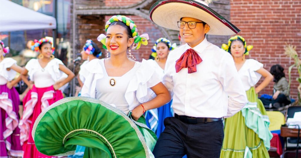 A pair of dancers leading a folklorico performance in bright traditional costumes; the woman holds a green skirt while the man wears a white shirt, red necktie and a wide hat, with other dancers in colorful dresses in the background.