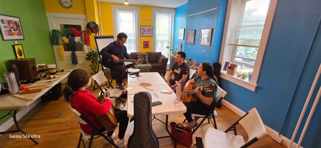Group of people in a colorful room playing guitars around a table, with a standing guitarist performing for others.