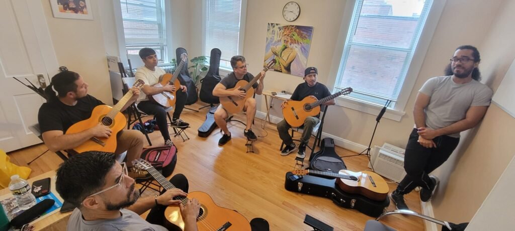 A casual guitar jam session in a bright room with six men seated and playing classical guitars while another man stands near the wall.