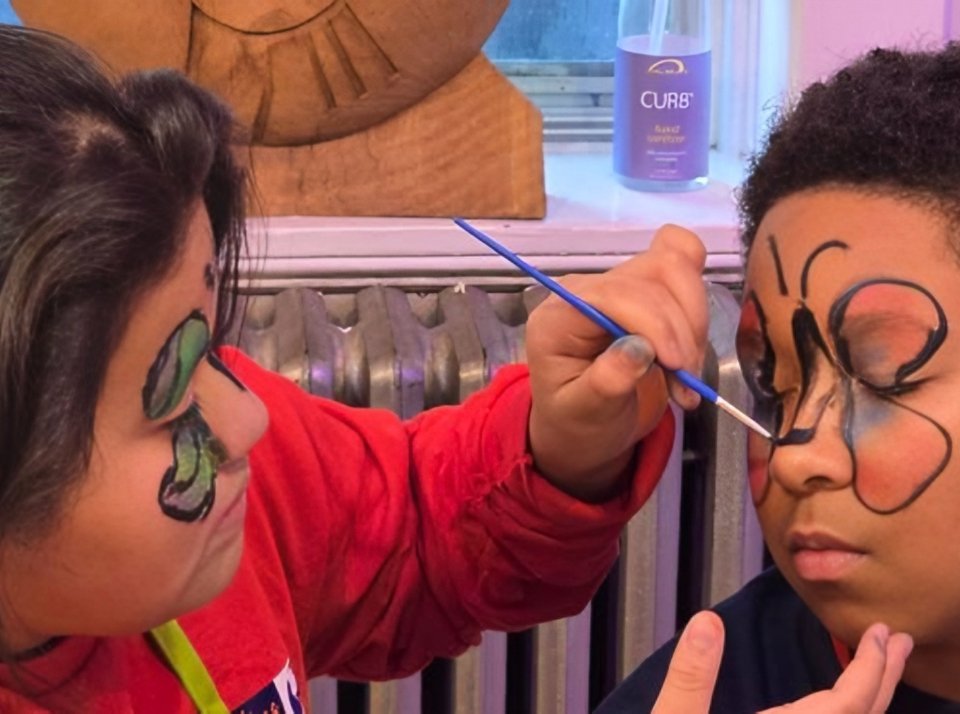 Two children at a face-paint station; one applies a black floral design around the other child's eye with a blue brush.